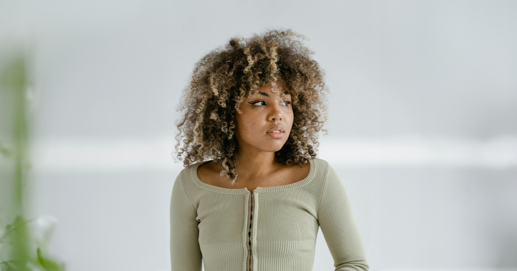 Young woman with curly hair wearing a light olive-green ribbed top, looking to the right in a bright, neutral space.